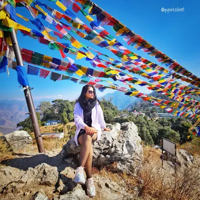 At the top of the world in Mussoorie, with Tibetan prayer flags fluttering in the wind. My travel styling focuses on layers, like this white jacket over a black dress, which are perfect for changing mountain weather.
