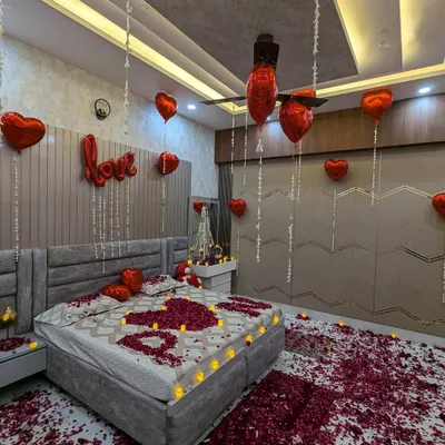 A classic romantic surprise setup. The room is filled with red heart balloons, a "LOVE" foil balloon, and a bed decorated with petals and LED candles spelling out "I ❤️ U".