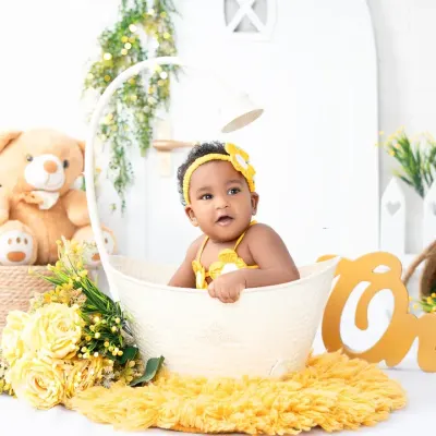 A sweet sitter session portrait of a baby girl in a bright yellow outfit, peeking out of a basket. These sessions are perfect for capturing babies once they can sit up on their own.