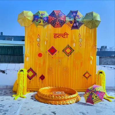 A vibrant and traditional Haldi ceremony setup on a rooftop. The decor is centered around a bright yellow backdrop, Rajasthani umbrellas, and marigold flower garlands.