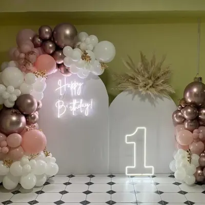 A chic and modern first birthday setup with white arched backdrops, a neon sign, a "1" marquee number, and a beautiful balloon arrangement in rose gold, pink, and white.