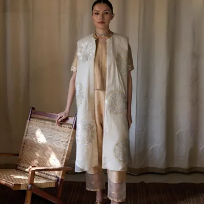 A full-length shot for Bellerobe, showcasing a layered ethnic ensemble. The natural light from the window highlights the delicate embroidery on the off-white jacket, while the simple cane chair adds a touch of organic texture to the scene.