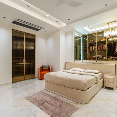 A wide view of a master bedroom that exudes luxury and tranquility. The design features a stunning walk-in closet with textured glass and gold-finished doors, which serves as a beautiful backdrop to the plush, neutral-toned bed.