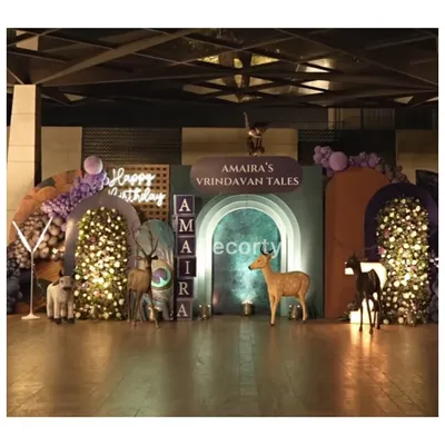 A mystical "Vrindavan Tales" birthday theme. This setup features enchanting backdrops with deer, cows, and floral arches, all illuminated to create a magical nighttime forest feel.