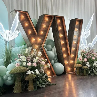 An enchanted forest themed decor featuring large rustic marquee letters. This whimsical setup includes pastel balloons, illuminated butterfly figures, and mossy mushroom props.