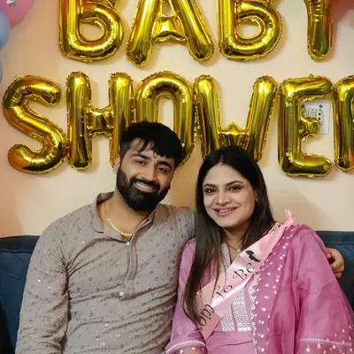A happy couple posing in front of a simple 'Baby Shower' decoration featuring large gold foil letters.