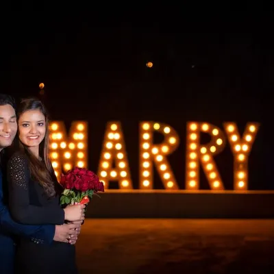 A bold and beautiful proposal setup for a pre-wedding shoot. The large, illuminated "MARRY ME" sign creates a stunning focal point for this nighttime portrait.