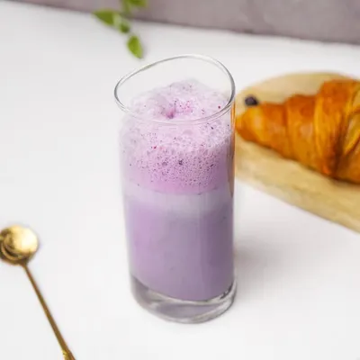 A bright blueberry smoothie in a tall glass, paired with a croissant in a clean, lifestyle cafe setting.