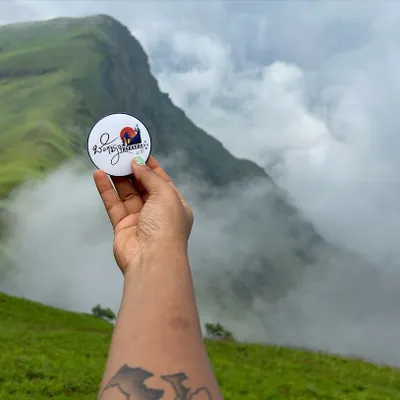 Our brand badge held up against the stunning, cloud-covered peaks of the Kudremukha range. This is what monsoon trekking in Karnataka is all about.