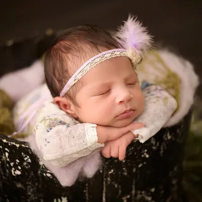 A classic newborn pose, with the baby sleeping peacefully in a dark bucket, wearing a delicate floral outfit and a lace headband.