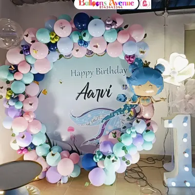 A beautiful mermaid-themed first birthday party for Aarvi. This setup features a circular backdrop with a custom design, a full balloon garland in ocean colors, and a large light-up number '1'.