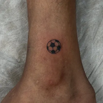 For the love of the game. A tiny, minimalist football tattoo on the ankle, created with clean lines and solid black shading. It's a small but powerful symbol of passion.