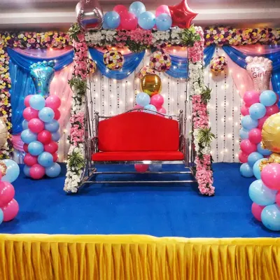 A fun and colorful baby shower stage for a baby girl. The setup includes pink and blue balloon pillars with baby-shaped foil balloons, a decorated swing, and a floral backdrop.