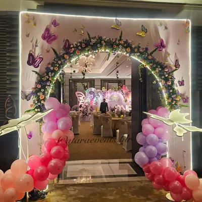 Welcome to a magical wonderland. This stunning entrance features a floral arch with butterfly accents, illuminated by soft lights and framed by pink and purple balloon garlands.