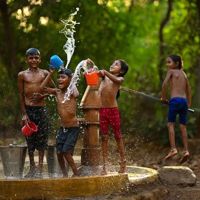 A girl leaps into the air with pure delight as her friends splash water around the village handpump. This action shot perfectly freezes the peak of summer fun and carefree energy.