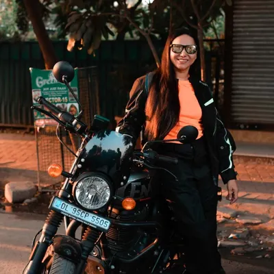 A confident female rider posing with her Royal Enfield Hunter. The warm evening light and urban setting create a cool, atmospheric portrait that captures the essence of modern motorcycling culture.