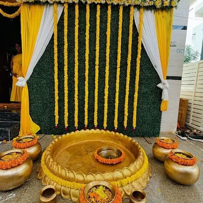 A traditional Haldi setup featuring a central brass urli for the ceremony, surrounded by smaller pots. The backdrop combines a lush green wall with vertical strings of marigold flowers and elegant yellow and white drapes.