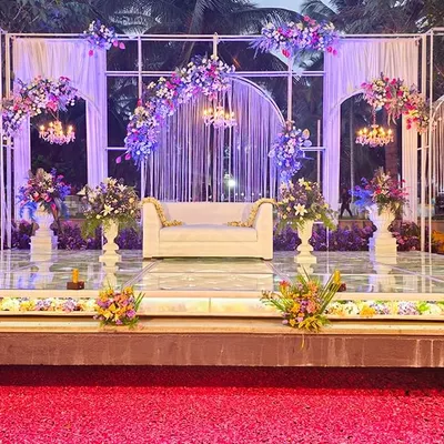A panoramic view of a glamorous outdoor reception stage. The design features elegant white arches, cascading string lights, and lush floral arrangements, with a pathway of red petals leading to the stage.