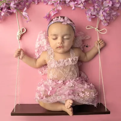 A little angel sleeping peacefully on a swing. This setup uses delicate props like the floral wings and a soft pink backdrop to create a dreamy, ethereal portrait.