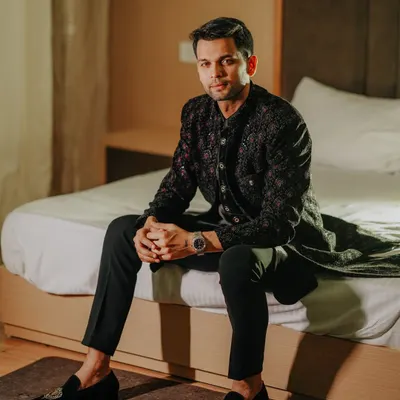 A handsome portrait of a groom dressed in a stylish black embroidered outfit, seated on a bed in a moment of quiet contemplation before the event.