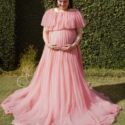 A beautiful full-length portrait taken in a sunlit garden. The long, flowing pink gown looks stunning against the lush green grass and foliage.