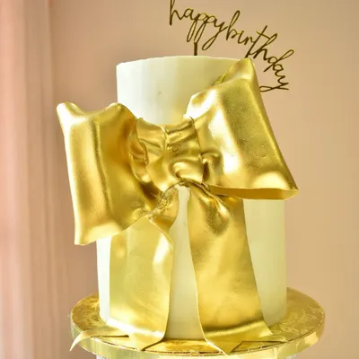 A statement of pure elegance. This two-tier white cake is wrapped in a large, luxurious, hand-sculpted golden bow, making it a glamorous centerpiece for a sophisticated birthday or anniversary celebration.