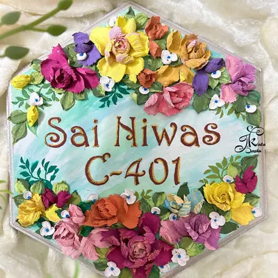 A beautiful hexagonal nameplate for "Sai Niwas," featuring a refreshing light blue base and a wreath of vibrant, multi-colored sculpted flowers. The brown lettering stands out beautifully against the background.