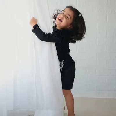 A joyful, candid shot of a little girl in a black outfit laughing as she plays with a white curtain.