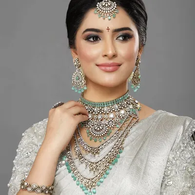 An editorial-style portrait featuring a stunning silver saree and layered diamond and mint-green jewelry. The makeup is clean and luminous, with a focus on flawless skin and a defined eye.