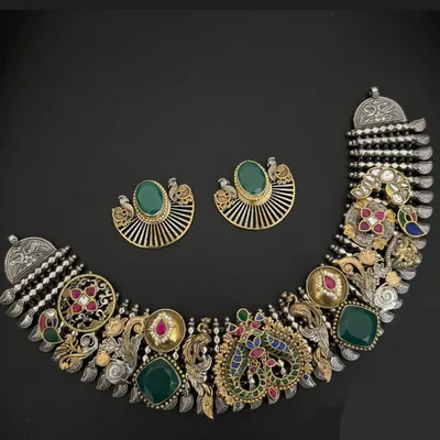 A masterpiece of fusion design. This statement necklace set combines oxidised silver with gold-plated elements, featuring intricate peacock motifs, large green stones, and detailed Kundan work for a truly royal look.