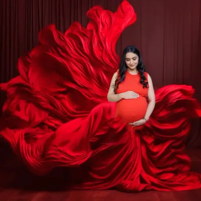 A full-body portrait of a mother-to-be in a magnificent red gown. The fabric is arranged to create a sense of movement and grandeur against a matching red backdrop.