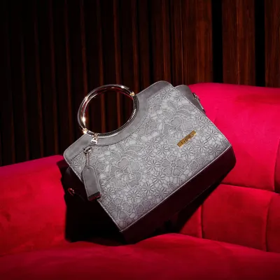 This elegant grey handbag with a unique metal ring handle is placed on a rich, red velvet couch. The luxurious setting and lighting highlight the bag's sophisticated design.