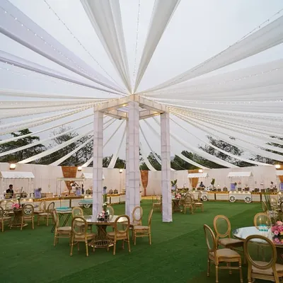 A wide-angle view of an outdoor event setup, featuring a magnificent white draped tent over lush green turf. This creates a bright and airy atmosphere for a daytime celebration.