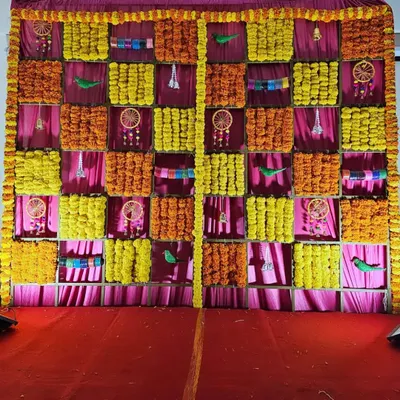 A traditional backdrop for a baby shower, featuring a checkerboard pattern of yellow and orange marigolds against a pink fabric. The small dreamcatchers and parrots add a unique, decorative touch.