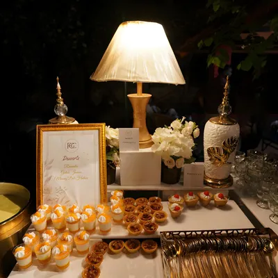 A beautifully lit dessert bar at an evening event. The table features a variety of treats, including mango dessert shots and mini tarts, arranged under a classic table lamp for a warm, inviting glow.