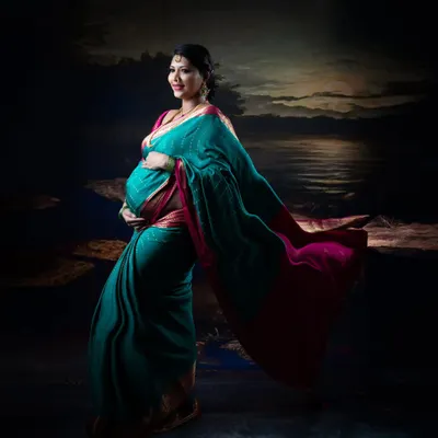An artistic maternity portrait of a woman in a teal and pink saree, with the fabric flowing beautifully against a painted seascape backdrop.