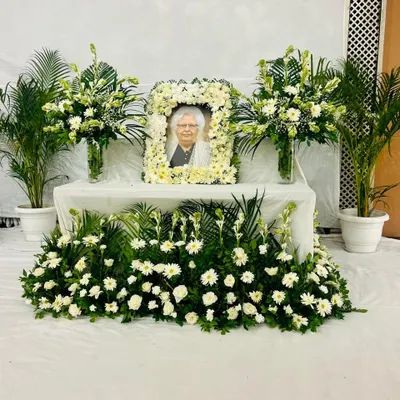 A full view of a memorial arrangement I delivered for a client in Noida. The setup includes a thick, handcrafted garland of white roses and chrysanthemums for the photo frame. It is complemented by tall glass vases holding gladiolus and tuberose, potted areca palms for a touch of green, and a dense base of white daisies and gerberas to complete the peaceful tribute.