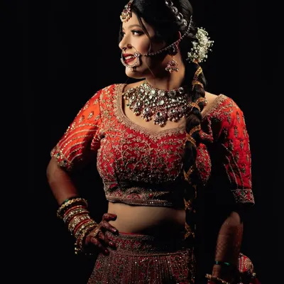 A striking bridal portrait against a dark background. The side profile and dramatic lighting highlight the intricate details of her red lehenga, jewelry, and confident pose.