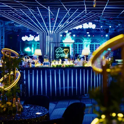 A chic and modern bar setup for a Sangeet night, featuring a "Gin O'Clock" neon sign and a facade of vertical blue LED lights that create a high-energy, club-like atmosphere.