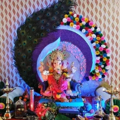 A unique Ganpati mandap featuring a large peacock theme. The backdrop is created with a fan of real peacock feathers and colorful paper flowers, with a sparkling circular frame for the Ganpati idol.