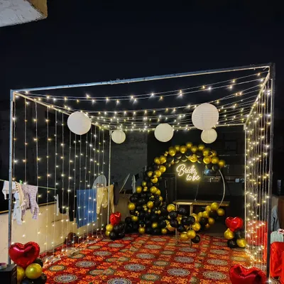 Celebrate the bride-to-be with a magical terrace party setup. This features a canopy frame draped in fairy lights, a black and gold balloon arch, and a 'Bride to be' neon sign for a chic and memorable night.