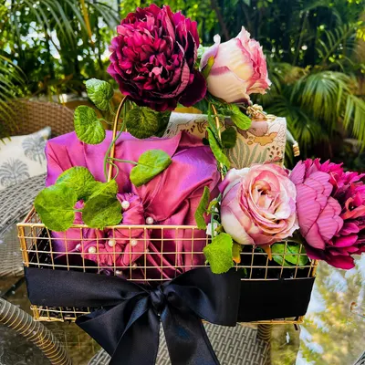 A wedding trousseau basket featuring gifts elegantly wrapped in vibrant magenta fabric. The arrangement is elevated with large, lush peonies and roses, all set within a chic gold-wired basket tied with a black satin bow.