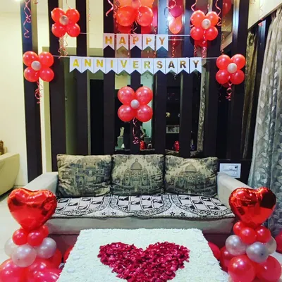 A simple and heartfelt anniversary decoration for a celebration at home. The setup includes red balloon arrangements, a "Happy Anniversary" banner, and a large heart made of rose petals on the table.
