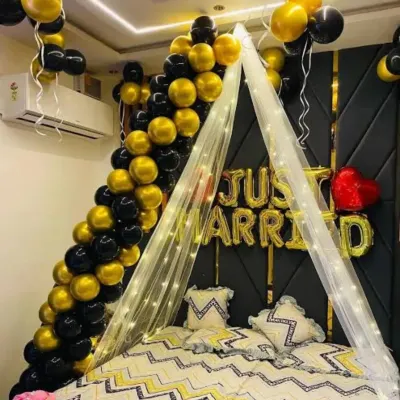 Welcome the newlyweds with a beautifully decorated room. This "Just Married" setup features a romantic canopy over the bed with fairy lights, complemented by a stylish black and gold balloon garland and foil letter balloons.