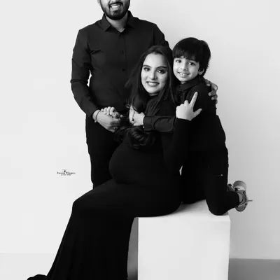 A joyful black and white family portrait. The mother-to-be is seated while her husband and son embrace her, all sharing a beautiful, happy moment.