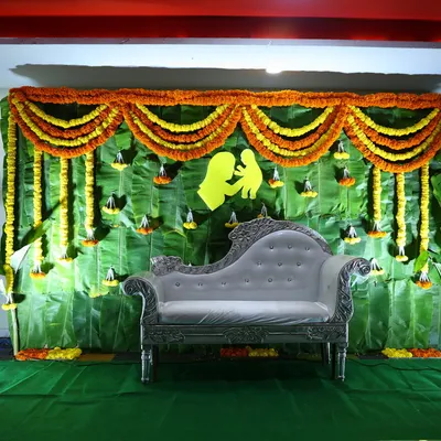 A traditional backdrop for a naming ceremony or baby shower, made entirely of fresh banana leaves and decorated with marigold garlands. A chaise lounge is placed for seating and photos.