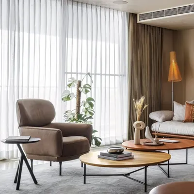 In the living area of Celadon, we layered textures and neutral tones to create a calm, inviting atmosphere. The soft brown armchair and nested wooden coffee tables sit before sheer curtains that diffuse natural light, enhancing the connection to the lush greenery just outside.