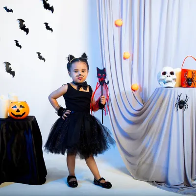 A child model in a full black cat Halloween costume, including a tutu, cat ears, and a wand, posing playfully in a spooky-themed set.