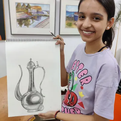 A student proudly shows her completed still life drawing of a metallic jug, demonstrating excellent skill in pencil shading to create realistic textures and reflections.