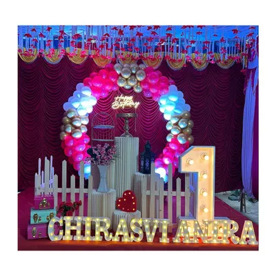 A festive first birthday stage with a circular balloon arch in red, white, and gold. The setup is personalized with the child's name in large marquee letters.